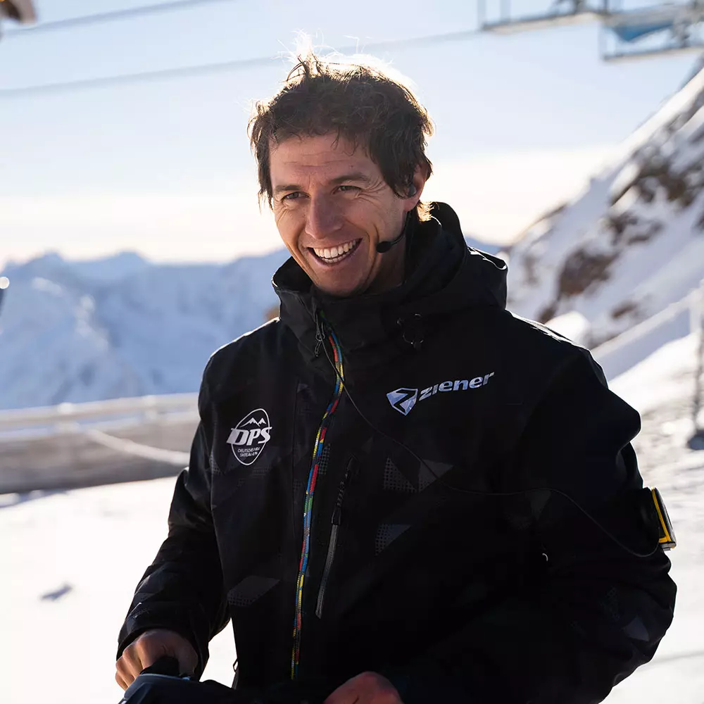 Benedikt Staubitzer_Headset lächelt vor verschneiten Bergen im Sonnenschein. Benedikt Staubitzer in Winterjacke mit Headset lächelt vor verschneiten Bergen im Sonnenschein.