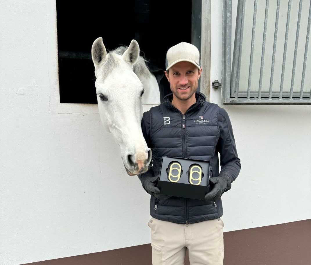 Christian Kukuk_hält eine schwarze Box mit zwei CEECOACH PLUS-Geräten, neben ihm ein weißes Pferd schaut aus einem Stallfenster Christian Kukuk in schwarzer Jacke und weißer Mütze hält eine schwarze Box mit zwei CEECOACH PLUS-Geräten, neben ihm ein weißes Pferd schaut aus einem Stallfenster