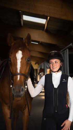 Eine Reiterin in Reitsportbekleidung, inklusive Helm und der Horse Pilot E-Twist'Air Airbagweste, steht neben einem braunen Pferd in einem Stall.