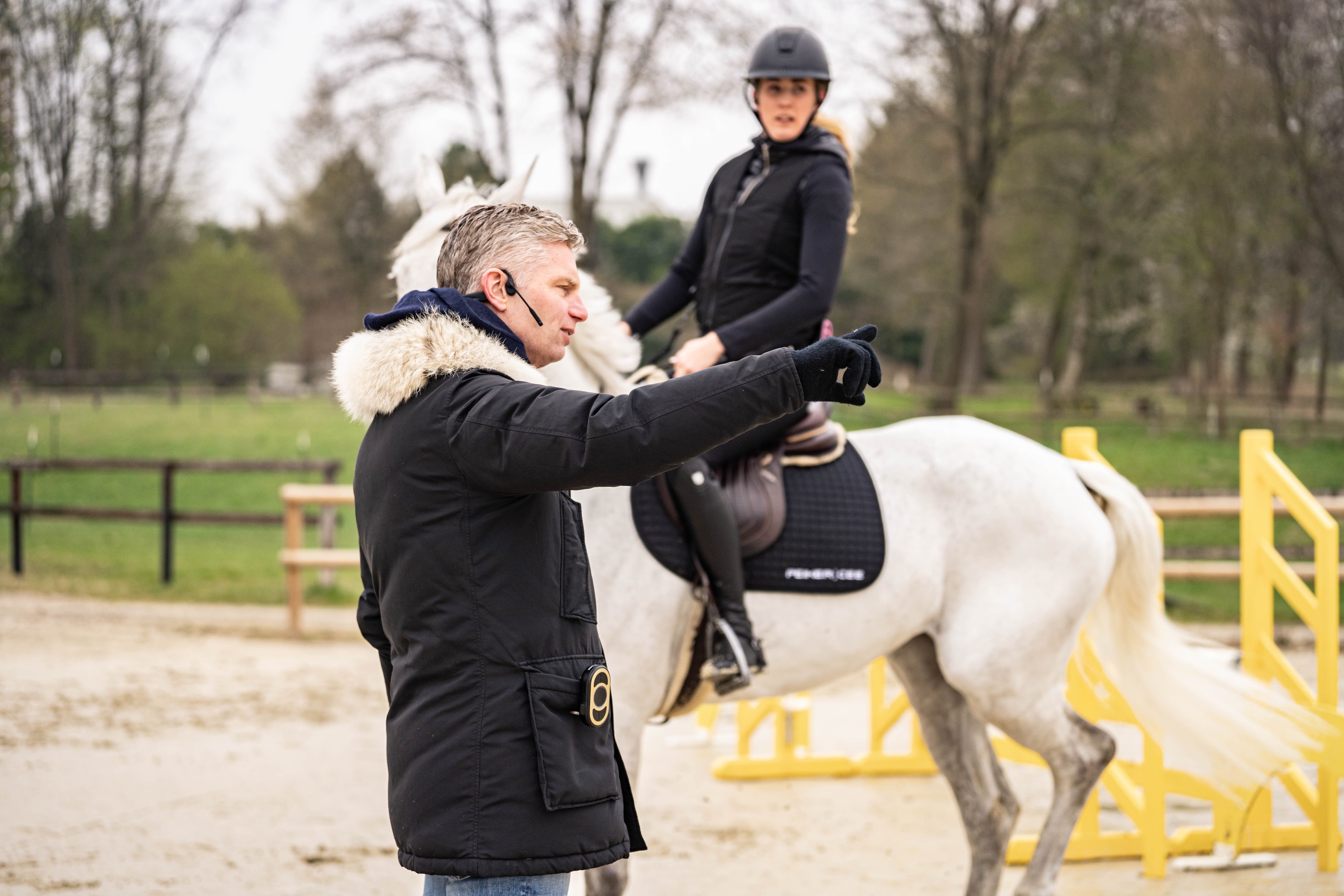 Reittrainer_steht im Vordergrund auf einem Reitplatz und zeigt mit ausgestrecktem Arm und Zeigefinger nach rechts_Im Hintergrund sitzt eine Frau einem weißen Pferd 