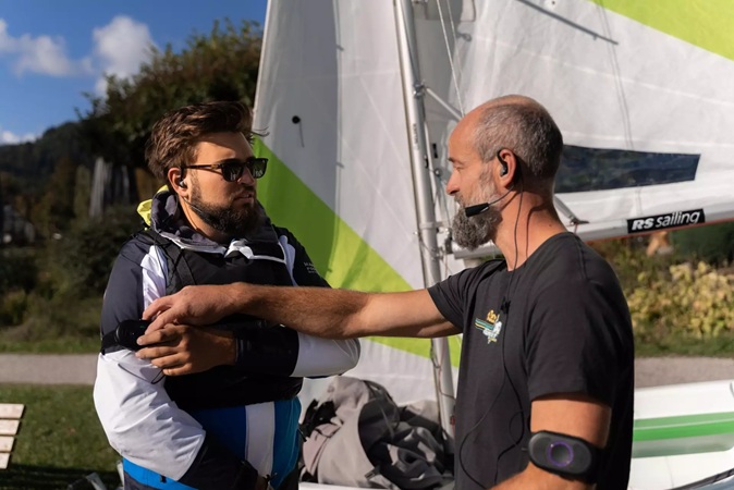 Segeltrainer erklärt einem Segler am Segelboot die Kommunikation mit dem CEECOACH PLUS, das er am Oberarm trägt.