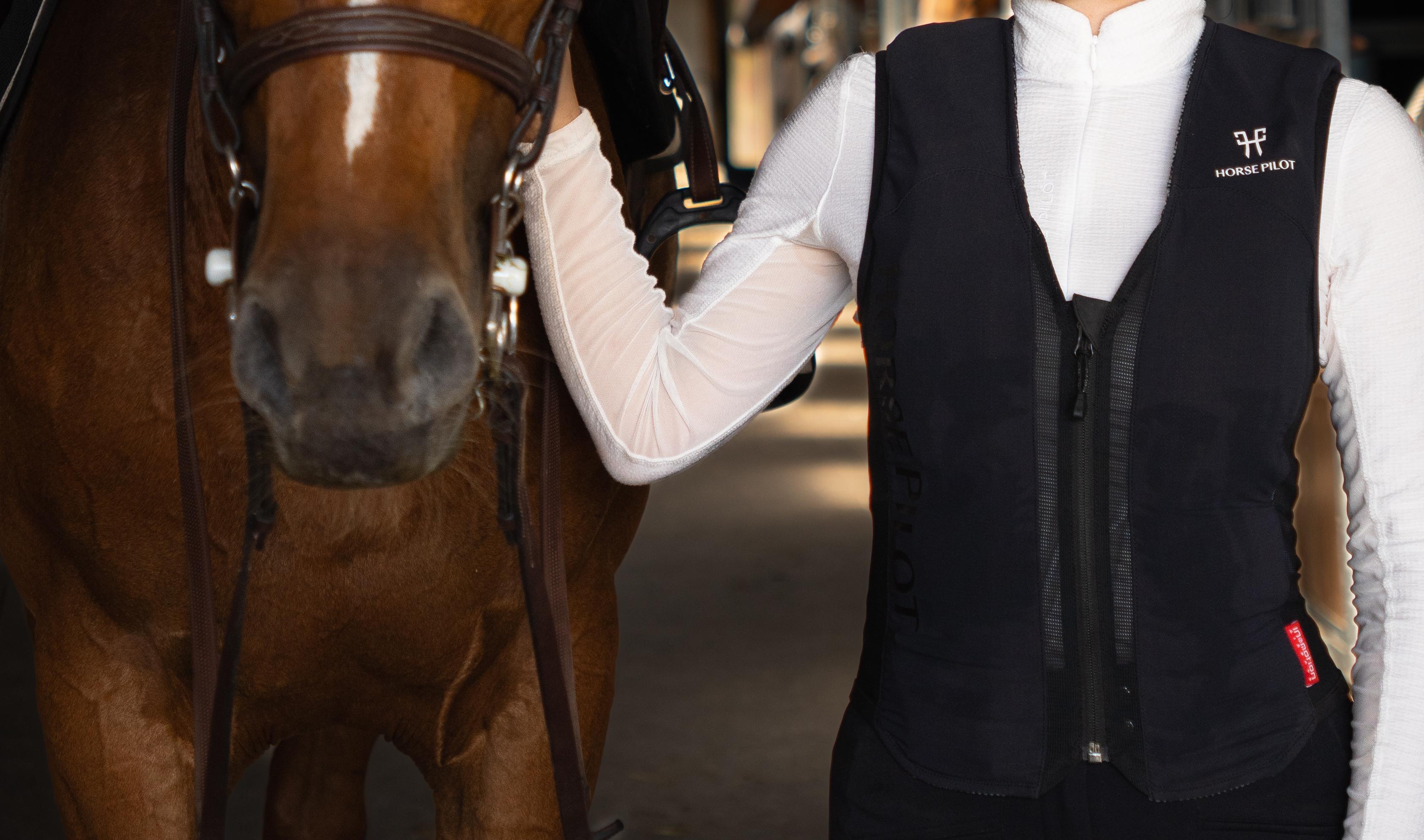 Nahaufnahme_Reiterin in Reitsportbekleidung, mit Helm und der Horse Pilot E-Twist'Air Airbagweste, steht neben einem braunen Pferd in einem Stall.
