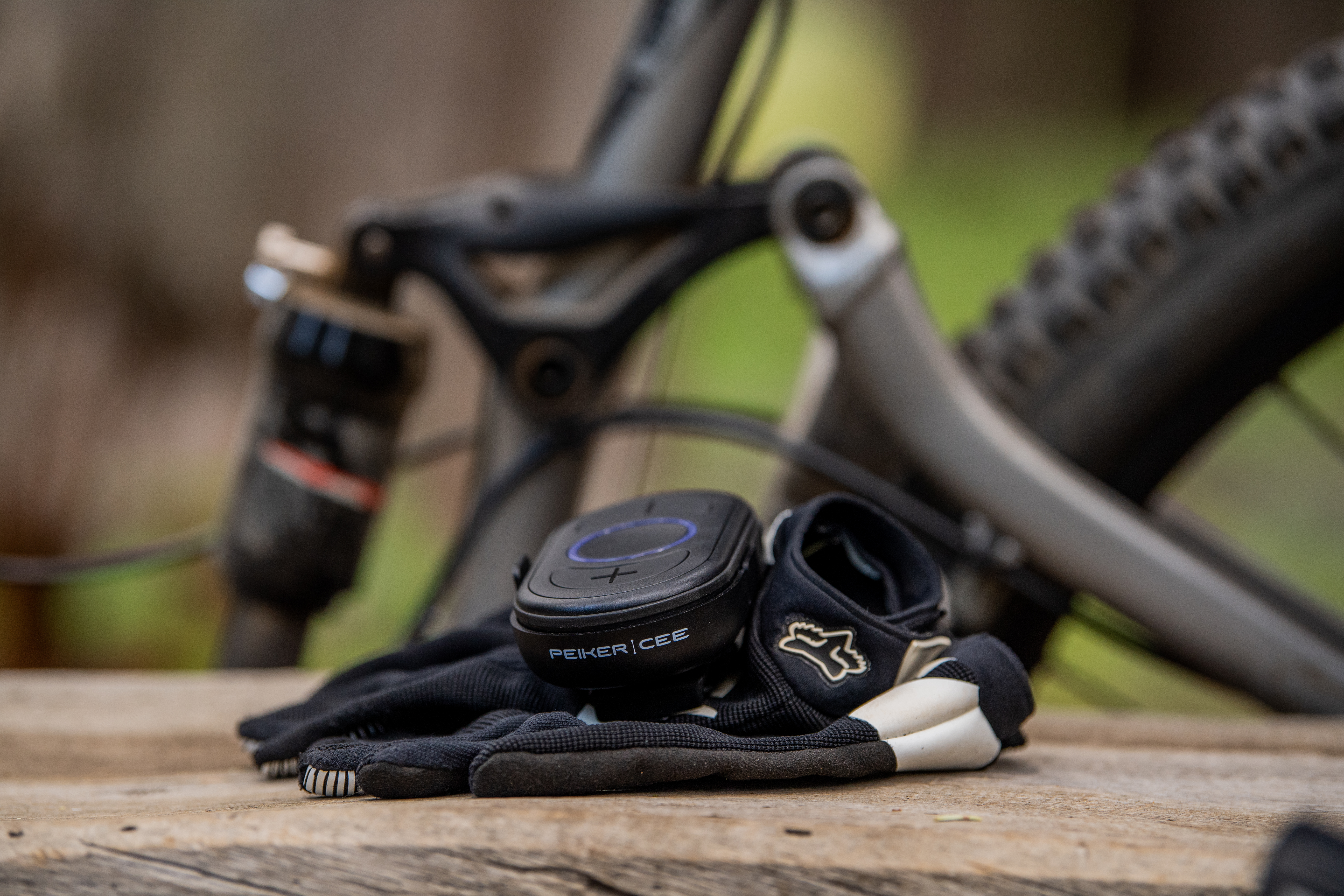 Ein CEECOACH PLUS liegt auf Handschuhen auf einer Holzoberfläche. Im Hintergrund ist unscharf ein Mountainbike zu sehen, was auf den Einsatz beim Outdoor-Training oder Radsport hinweist.