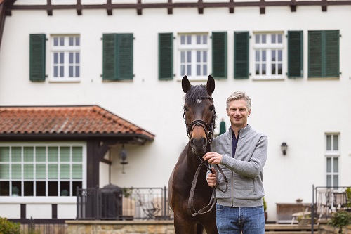 Lars Meyer zu Bexten stands with his horse in front of a white house and smiles at the camera.