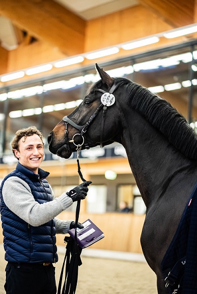 Calvin Böckmann smiles at the camera next to a horse in an indoor riding arena.