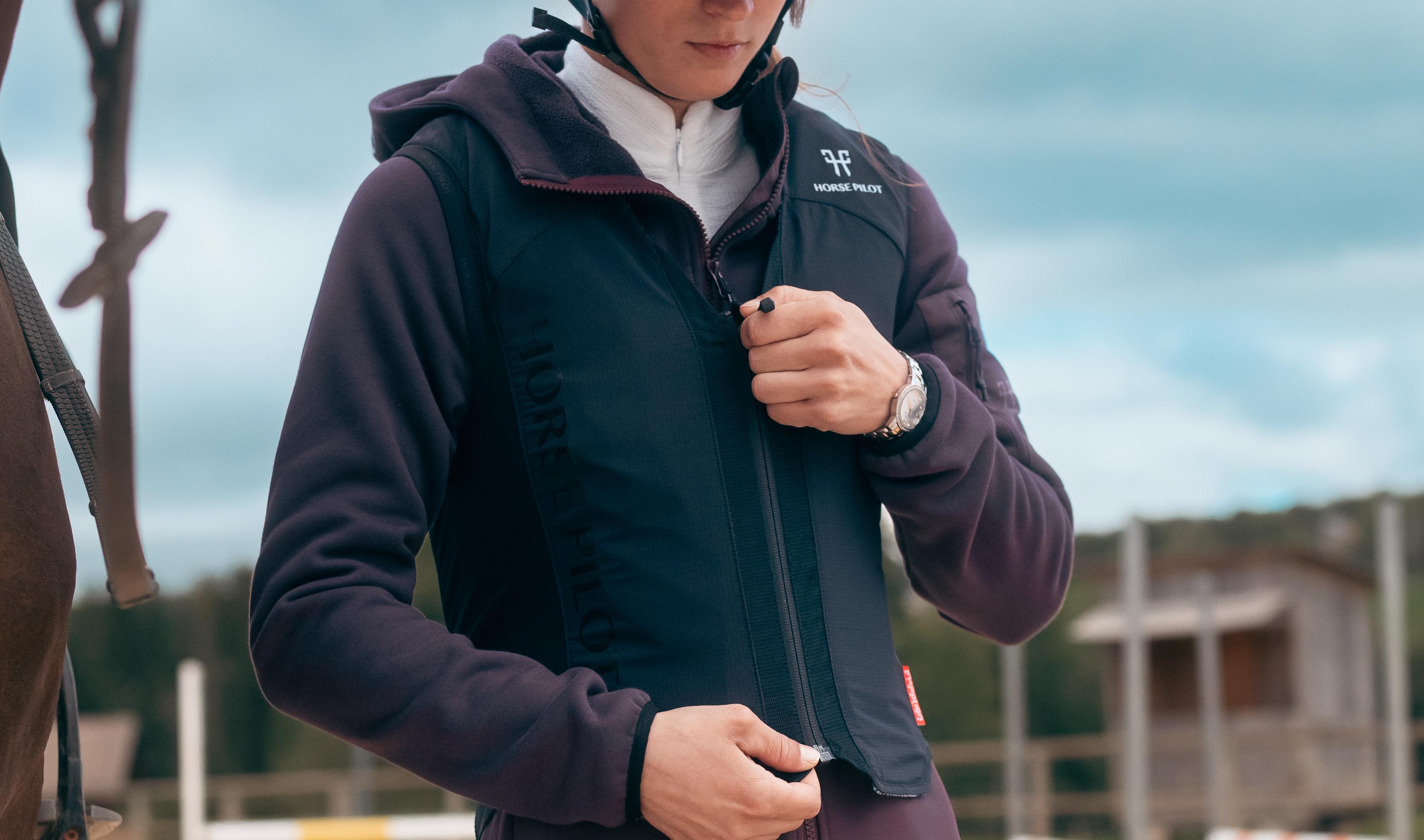 Reiter zieht sich die Horse Pilot E-Twist'Air Airbag-Weste an. Ihre Hände sind am Reißverschluss, während sie die Weste schließt. Im Hintergrund ist ein Reitplatz mit Hindernissen zu erkennen.